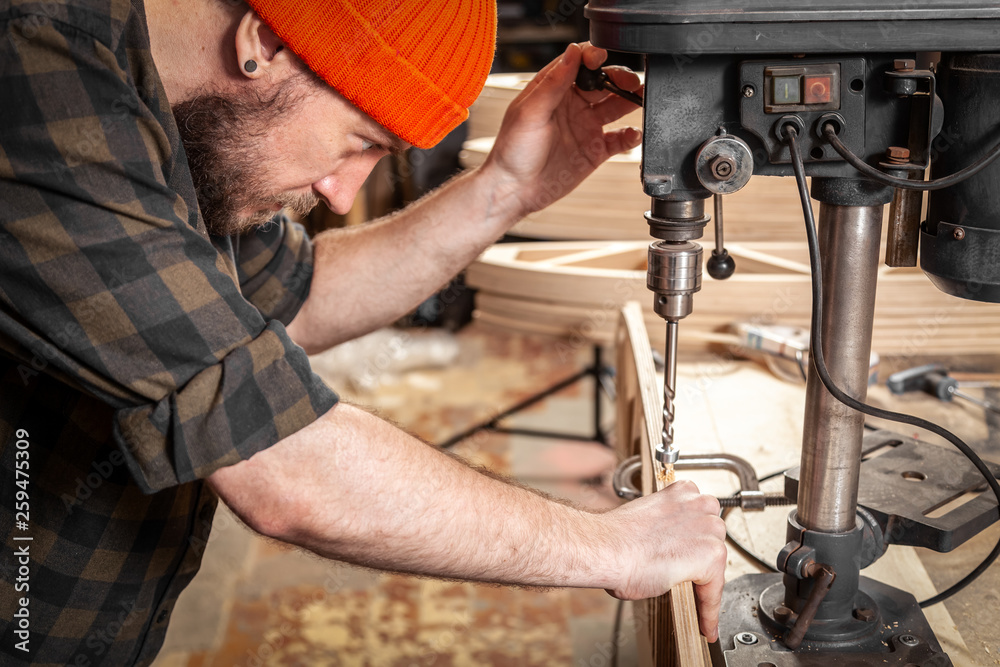 Close up of a man with work clothes and a carpenter's cap is carving a wooden board on an modern large drilling machine in a light workshop. Home repair concepts Close up of a man with work clothes and a carpenter's cap is carving a wooden board on an modern large drilling machine in a light workshop. Home repair concepts