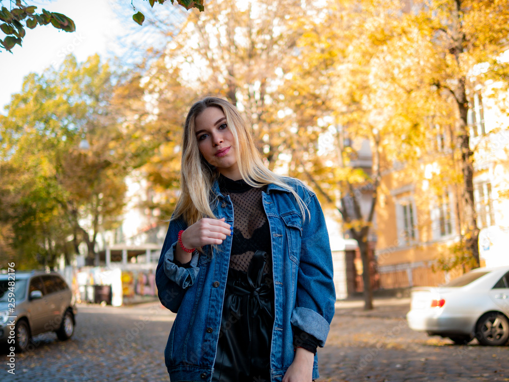 cute blonde girl with flowing hair in a jeans jacket standing on the street against the background of yellow trees