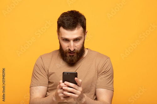 Close up shot portrait of young bearded man isolated on yellow background, looking agitated at display of her smartphone smiling and laughing happily, impressed by media content from web