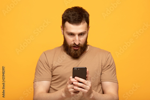 Close up shot portrait of young bearded man isolated on yellow background, looking agitated at display of her smartphone smiling and laughing happily, impressed by media content from web