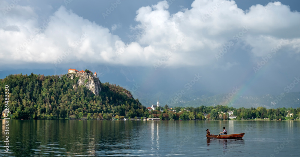 Amazing photo of Lake Bled at evening after rain with vibrant rainbow on the sky and couple in wooden boat. Slovenia