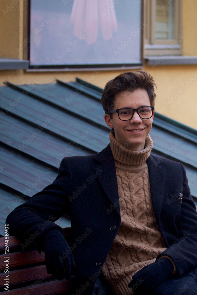 A young man with a smile sitting on a bench. Dressed in a black light overcoat, black-rimmed glasses. A young man with a smile sitting on a bench. Dressed in a black light overcoat, black-rimmed glasses.