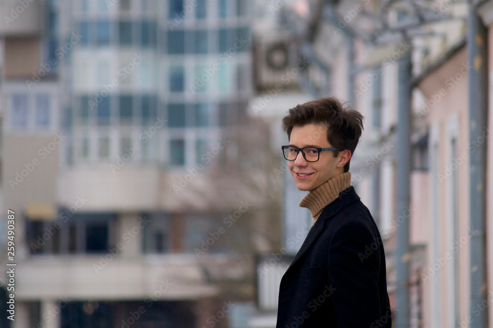 A young man posing with a smile, among the cityscape. Dressed in a black light overcoat, black-rimmed glasses. A young man posing with a smile, among the cityscape. Dressed in a black light overcoat, black-rimmed glasses.