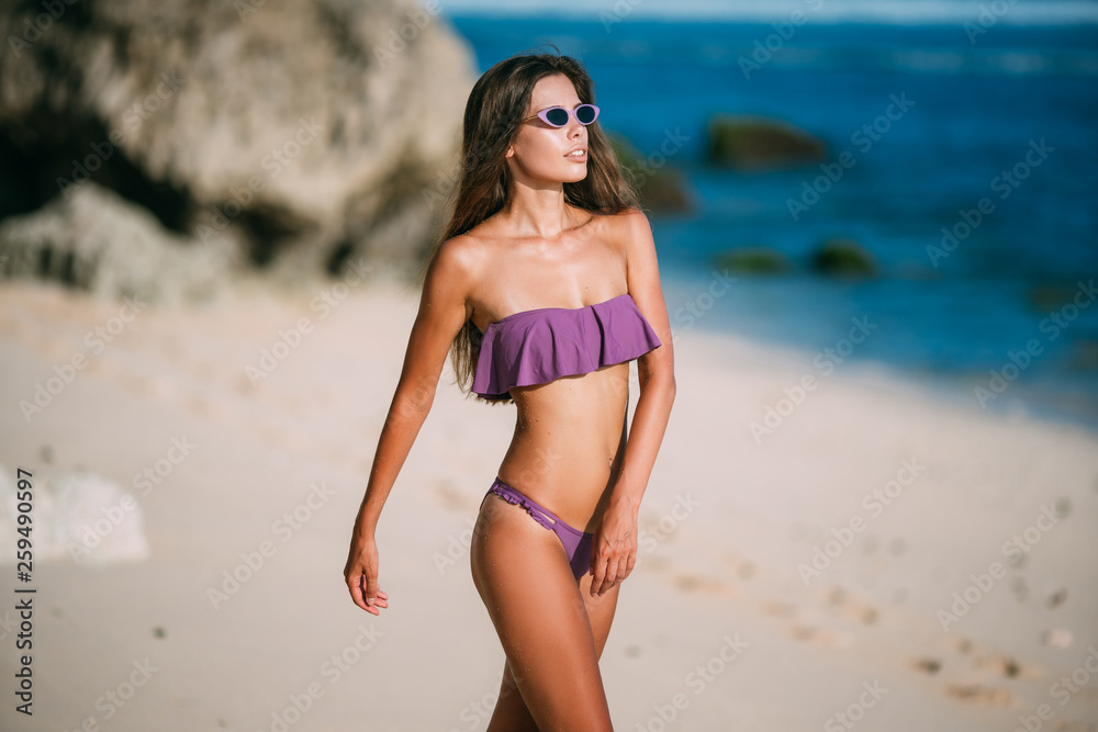 Slender sexy tanned girl in swimsuit posing on beach with sand and large stones Slender sexy tanned girl in swimsuit posing on beach with sand and large stones