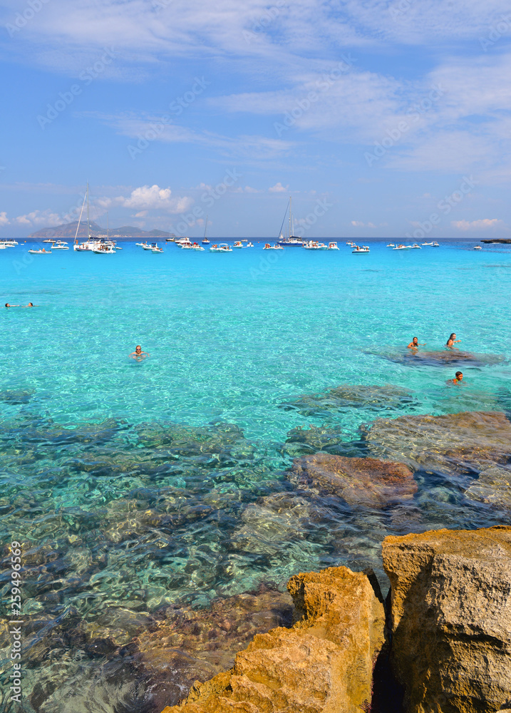 people inside paradise clear torquoise blue water with boats and cloudy blue sky in background in Favignana island, Cala Rossa Beach, Sicily South Italy.