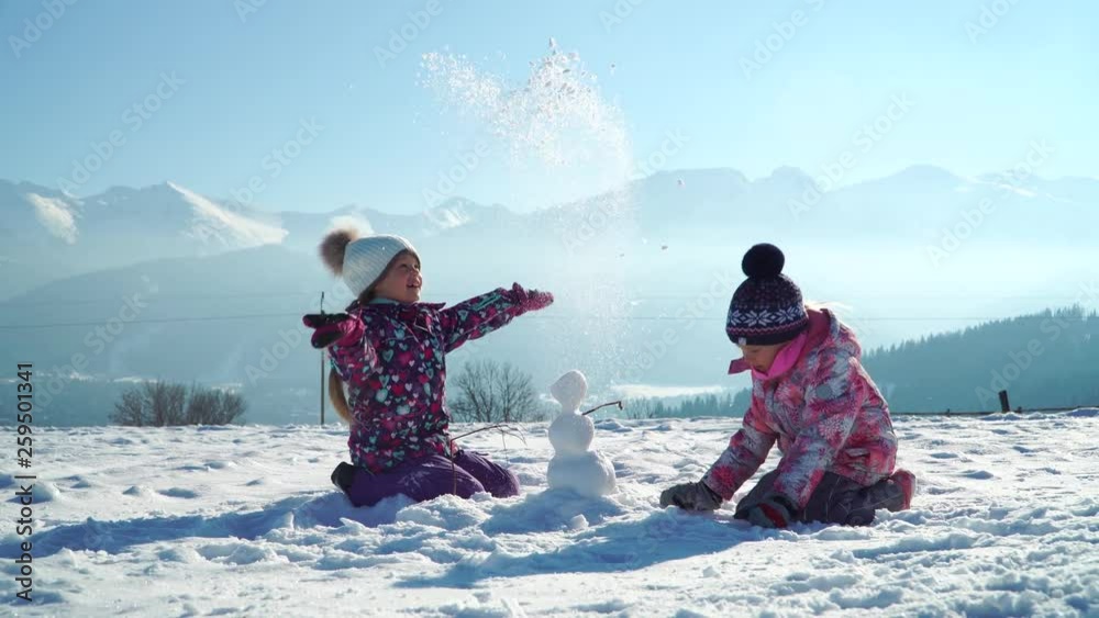 Children in outwear making small snowman while playing on snowy field in sunlight with mountains on background