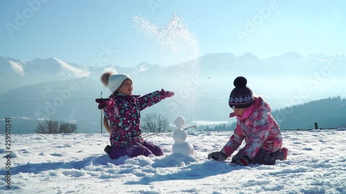 Children in outwear making small snowman while playing on snowy field in sunlight with mountains on background