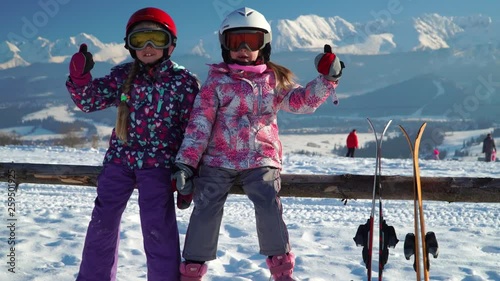 Little girls in outwear and masks with helmet sitting on wood fence with skis near against mountains
