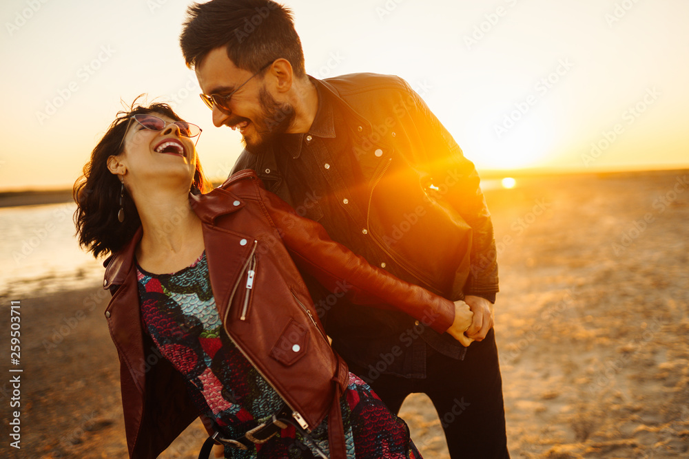 Enjoying time together.  Stylish and loving couple enjoying each other by the sea. The couple is young and in love. The concept of youth, love and lifestyle. Beautiful sunset on a summer day. - obrazy, fototapety, plakaty 
