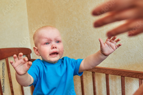 The little boy is crying in the crib and pulling his hands to the mom