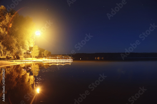 Lake or river sand shore with trees and glowing lantern over pantoon pier and dark blue starry sky on background. Tranquil nature night landscape