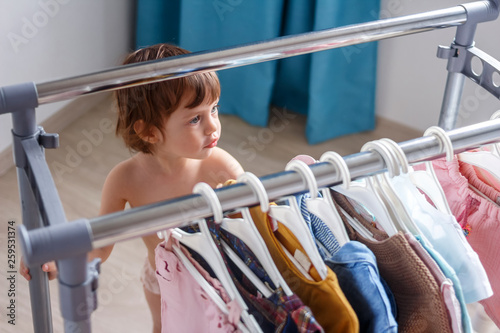 Pretty little girl fashionista with gray eyes and short hair chooses an outfit from trendy clothes hung on a rail. Soft Focus of a Two Years Old Child Choosing her own Dresses from Kids Cloth Rack.