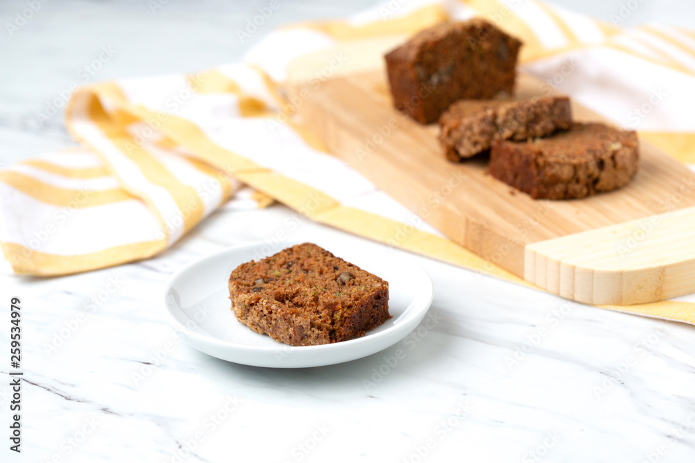 Homemade Zucchini Bread Sliced on Cutting Board with One Slice Isolated in Front on Small White Plate; Yellow and White Dish Towel on White Countertop Homemade Zucchini Bread Sliced on Cutting Board with One Slice Isolated in Front on Small White Plate; Yellow and White Dish Towel on White Countertop