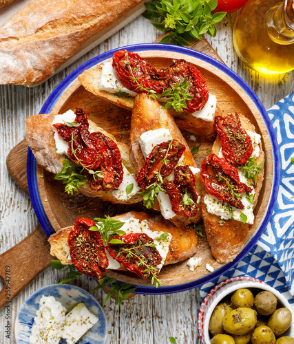 Bruschetta with feta cheese, dried tomatoes, olive oil and fresh aromatic herbs, on a plate on a wooden table, top view.  Delicious Mediterranean vegetarian appetizer