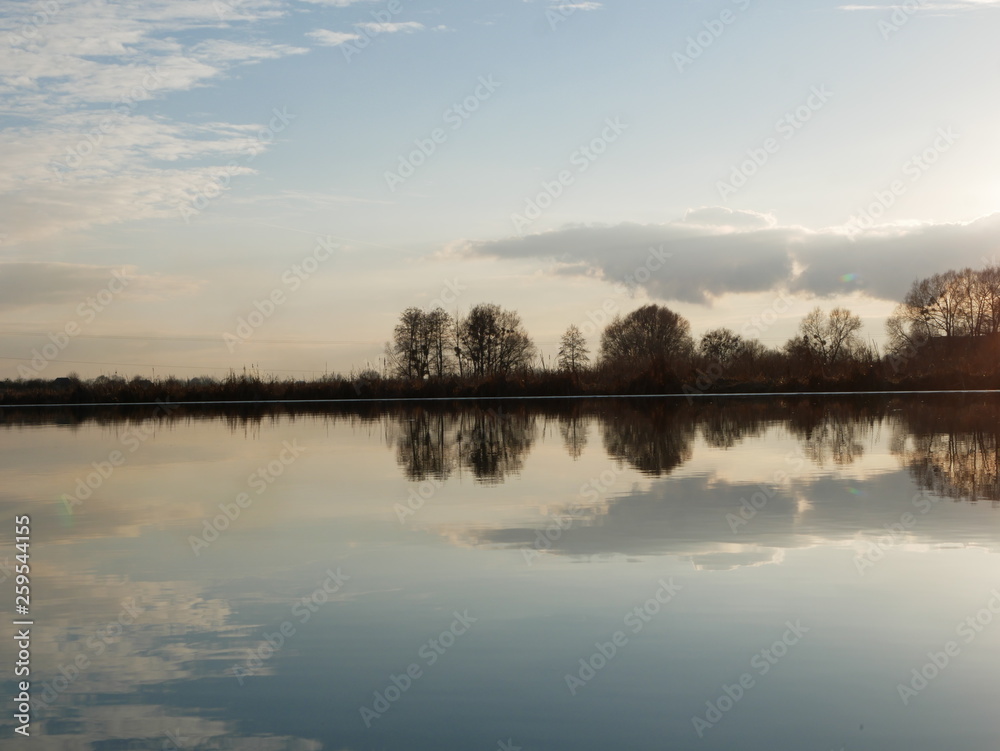 Polonne / Ukraine - 18 February 2019: Scenic river reflections of trees on the water.  A river with trees on the shores. Reflection of blue sky with clouds in calm water