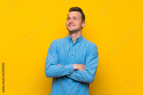 Blonde man over isolated yellow wall looking up while smiling