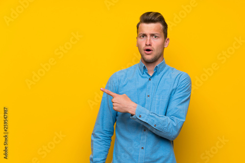 Blonde man over isolated yellow wall surprised and pointing side