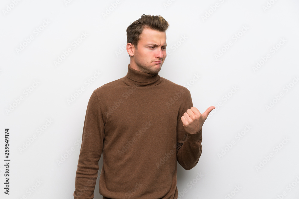 Blonde man over isolated white wall unhappy and pointing to the side Blonde man over isolated white wall unhappy and pointing to the side