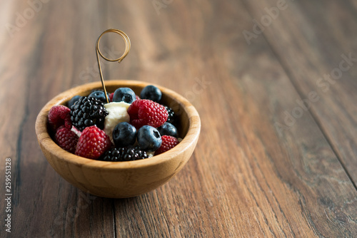 Delicious sweet dessert made from berry mix (raspberries, blueberries and blackberries) with whipped cream in a wooden bowl on a wooden background. Close up.