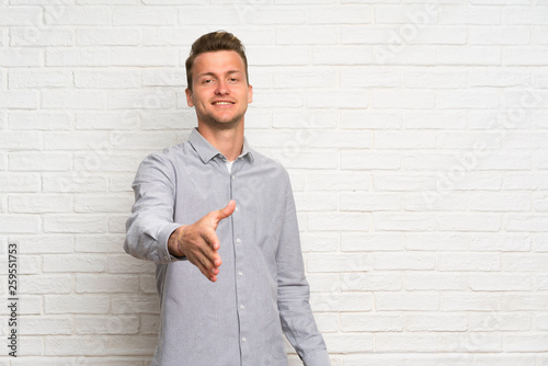 Blonde man over white brick wall shaking hands for closing a good deal