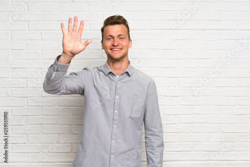 Blonde man over white brick wall counting five with fingers