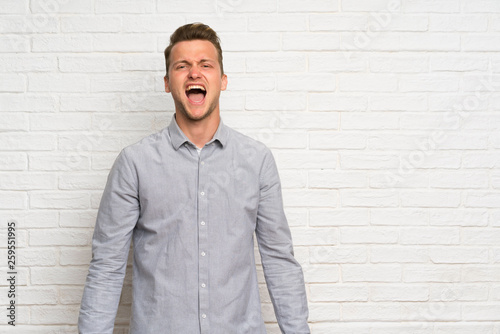 Blonde man over white brick wall shouting to the front with mouth wide open