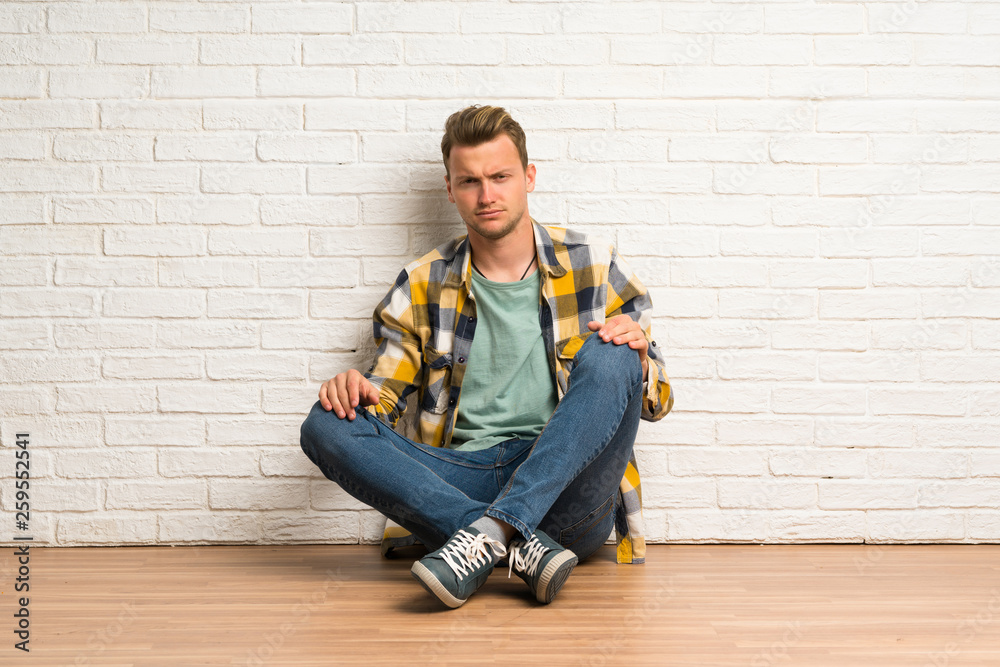 Blonde man sitting on the floor with sad and depressed expression Blonde man sitting on the floor with sad and depressed expression