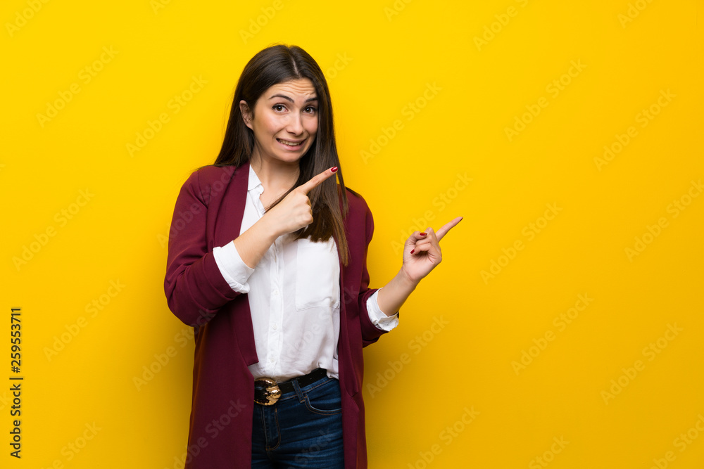 Young woman over yellow wall frightened and pointing to the side Young woman over yellow wall frightened and pointing to the side