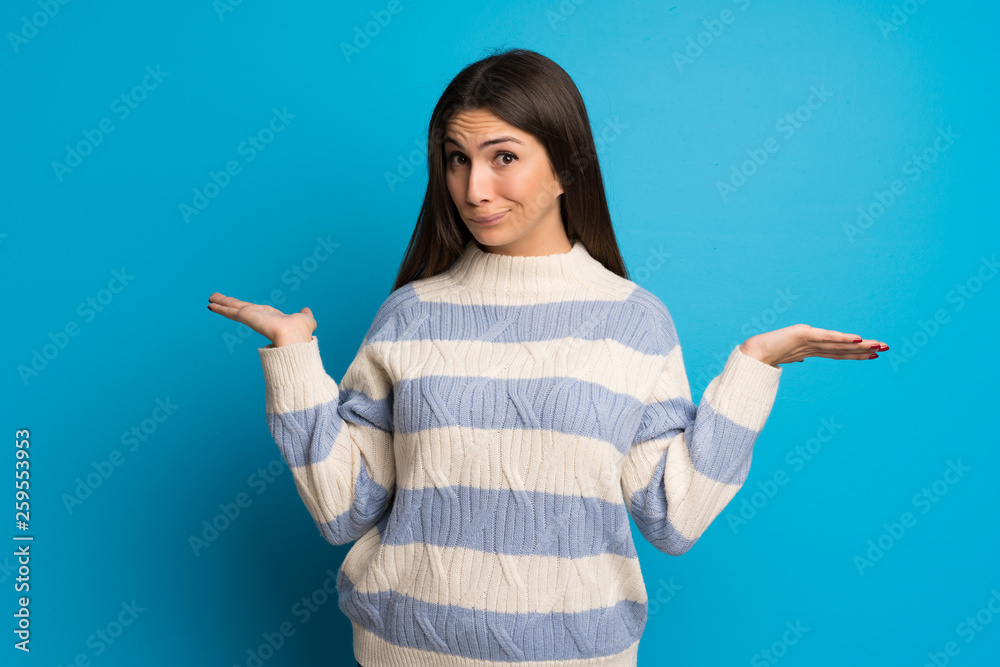 Young woman over blue wall having doubts while raising hands and shoulders Young woman over blue wall having doubts while raising hands and shoulders