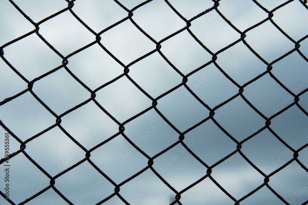 Metal black fence-mesh netting. Gloomy cloudy gray sky. The background image of the fence. Metal black fence-mesh netting. Gloomy cloudy gray sky. The background image of the fence.