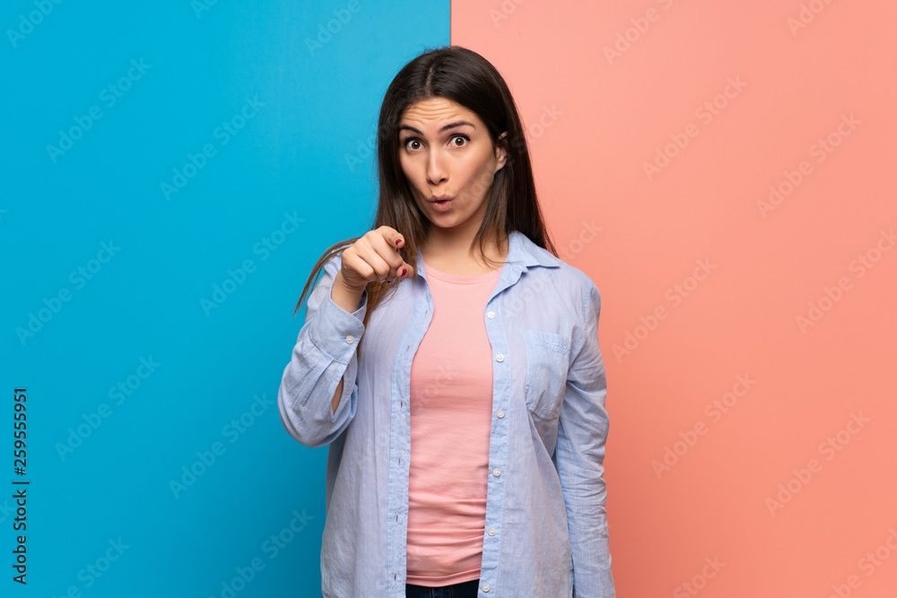 Young woman over pink and blue wall surprised and pointing front Young woman over pink and blue wall surprised and pointing front