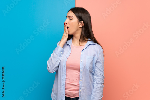 Young woman over pink and blue wall yawning and covering wide open mouth with hand © luismolinero