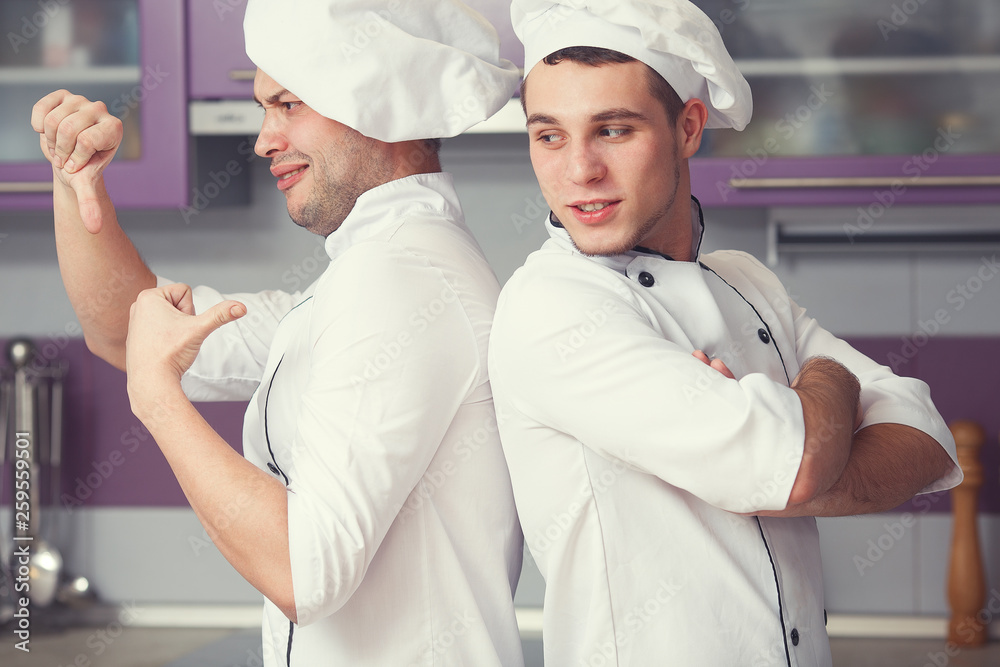 Portrait of two funny working men in cook uniform showing dislike sign (thumbs down) and posing in modern kitchen together. Close up. Indoor shot