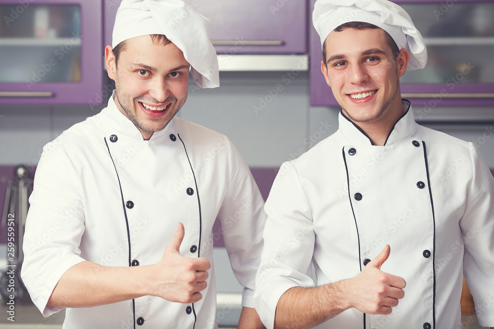 Portrait of two funny working men in cook uniform showing OK sign (thumbs up) and posing in modern kitchen together. Close up. Indoor shot