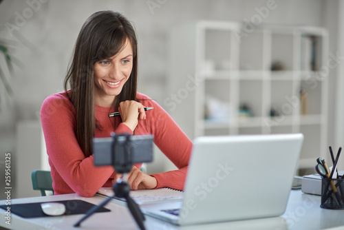 Charming Caucasian businesswoman sitting in modern office and taking notes in notebook. In front of her laptop.