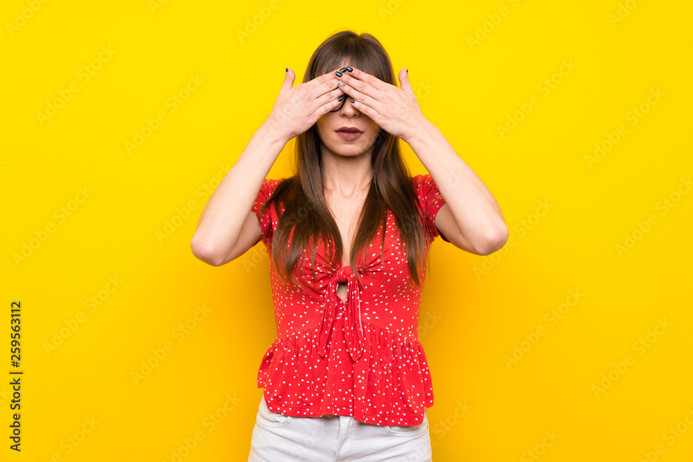 Young woman over yellow wall covering eyes by hands