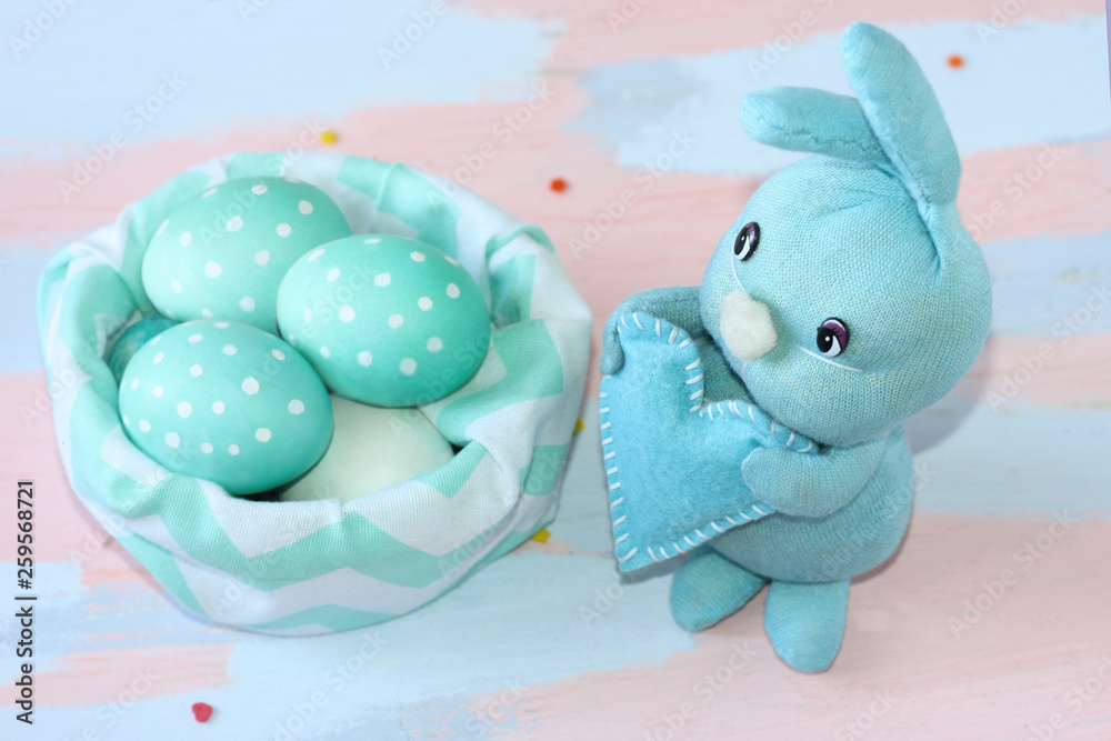 Easter light blue rabbit sewn by hands near a cloth basket with blue-white eggs . Handwork. Greeting card for happy easter. Easter light blue rabbit sewn by hands near a cloth basket with blue-white eggs . Handwork. Greeting card for happy easter.