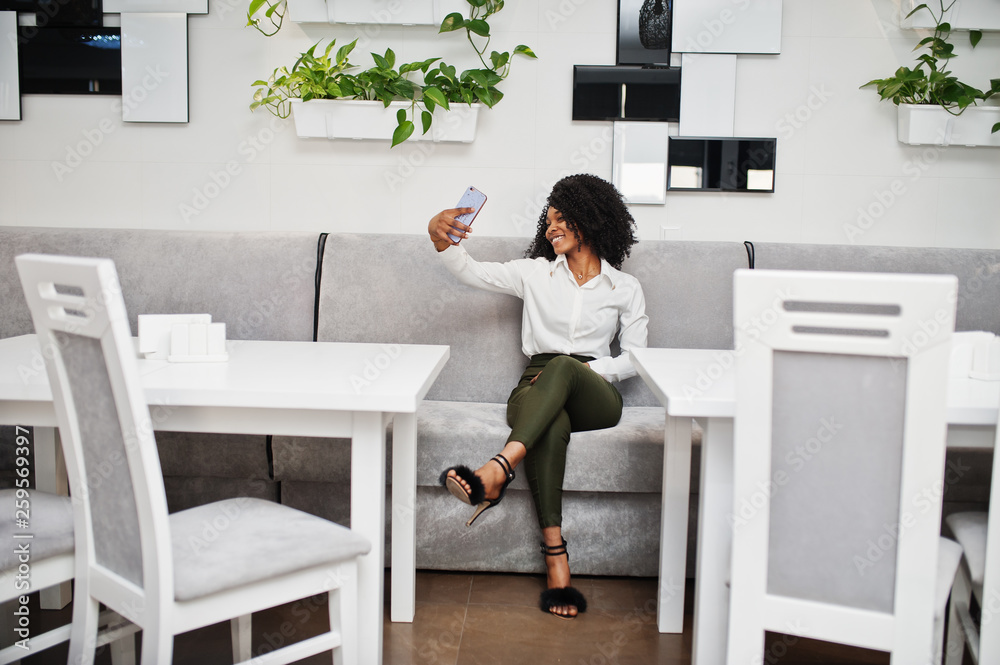 Сheerful business african american lady with afro hair, wear white blouse and green pants posed in cafe making selfie by cell phone.