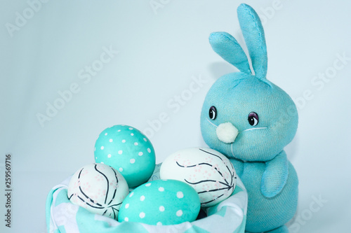 Easter light blue rabbit stands near the basket with blue-white eggs. Handmade. Greeting card for happy easter.