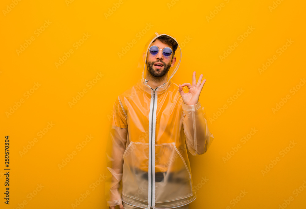 Young man wearing a rain coat cheerful and confident doing ok gesture Young man wearing a rain coat cheerful and confident doing ok gesture