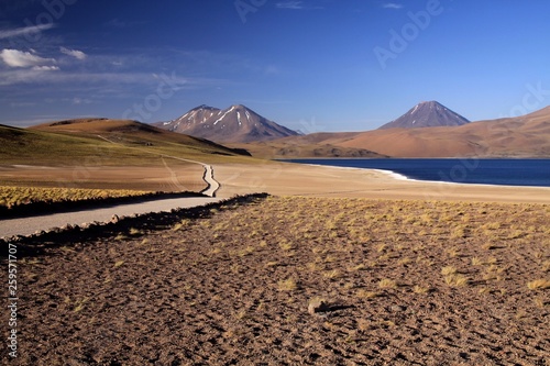 View on track along deep blue lake at Altiplanic Laguna (Lagoon) Miscanti in Atacama desert with partly snow capped cones of volcano Meniques in the background - Chile