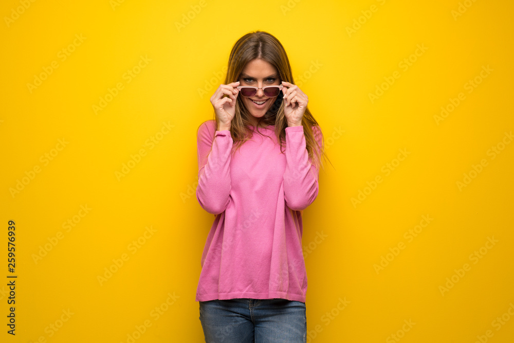 Woman with pink sweater over yellow wall with glasses and surprised Woman with pink sweater over yellow wall with glasses and surprised