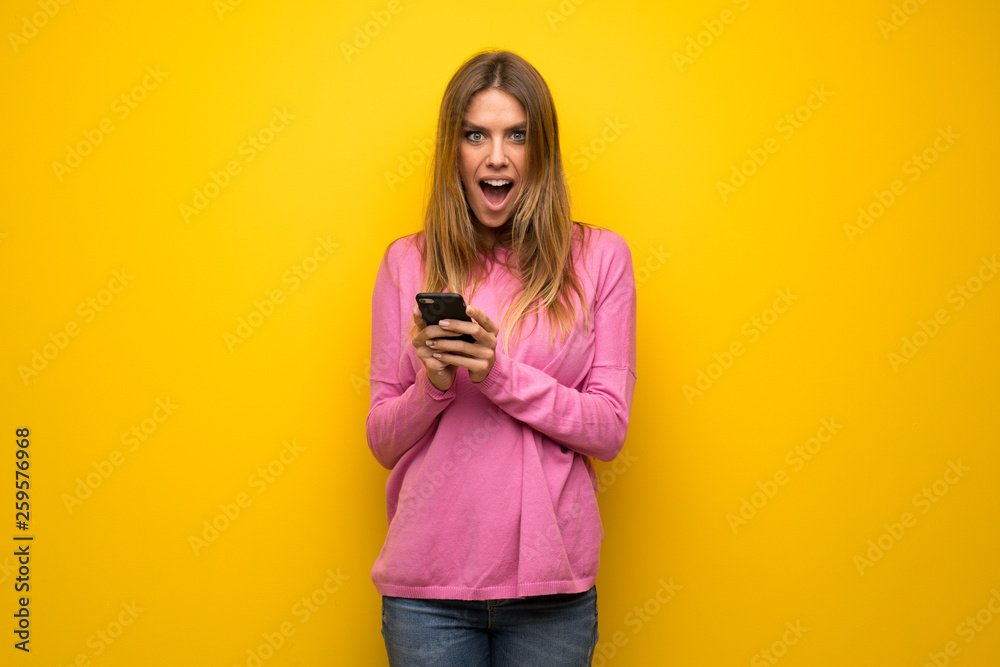 Woman with pink sweater over yellow wall surprised and sending a message Woman with pink sweater over yellow wall surprised and sending a message