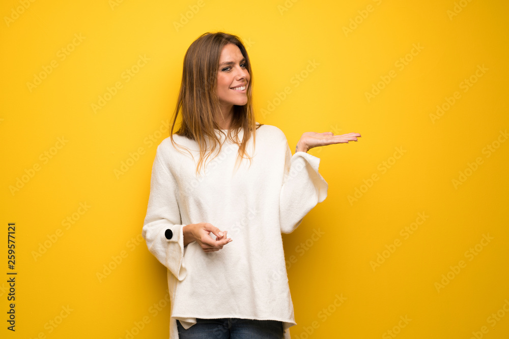 Blonde woman over yellow wall presenting an idea while looking smiling towards Blonde woman over yellow wall presenting an idea while looking smiling towards