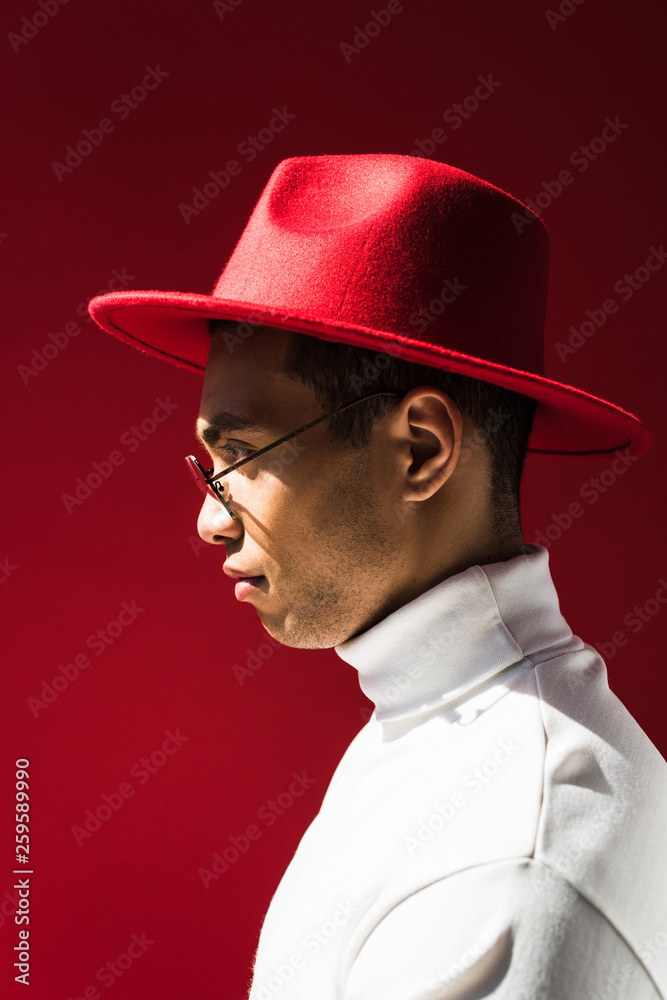 side view of stylish mixed race man in hat and glasses posing isolated on red side view of stylish mixed race man in hat and glasses posing isolated on red