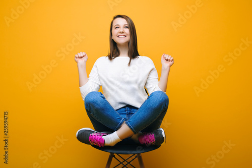 Portrait of a attractive caucasian woman sitting on a chair with crossed legs and looking up smiling white hands up because winning against yellow background.. Winning concept. photo