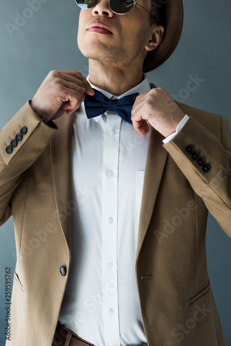 partial view of stylish mixed race man in suit adjusting bow tie isolated on grey