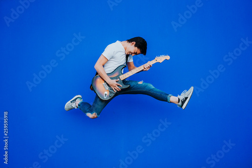 young man jumping with electric guitar on blue background