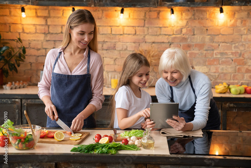 Cute little girl showing some to her mother and grandmother in the tablet while preparing vegetables in kitchen with light bulb on the background