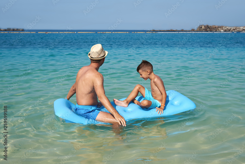 Attractive European man with his son is sitting on a blue air mattress on the beach. They are relaxing and enjoying their holidays.